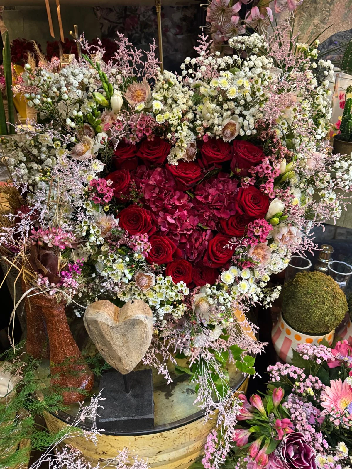 Herz aus roten Rosen und bordeauxfarbenen Blüten, umrahmt von zarten weißen und rosa Blumen